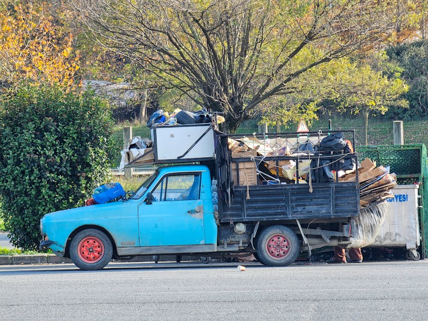 A small, older model light blue pickup truck parked on the side of a street during daylight hours. The vehicle's body shows signs of wear, with visible rust on the wheels and some dents on the door pa