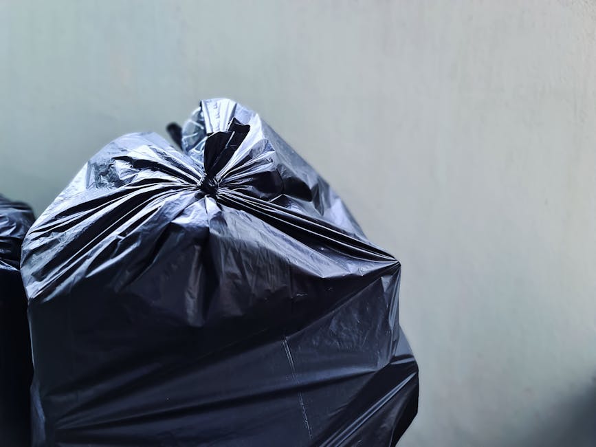 Close-up view of a tightly tied black plastic rubbish bag with a glossy, slightly crinkled texture, placed in the foreground against a plain, light-colored wall. The bag appears full and is positioned