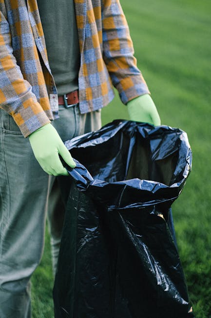 Close-up view of a tightly tied black plastic rubbish bag with a glossy, slightly crinkled texture, placed in the foreground against a plain, light-colored wall. The bag appears full and is positioned